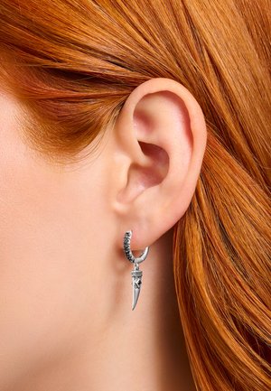 Nahaufnahme eines rothaarigen Ohrs, das einen silbernen Creolen-Ohrring mit einem detaillierten dolchförmigen Anhänger trägt.