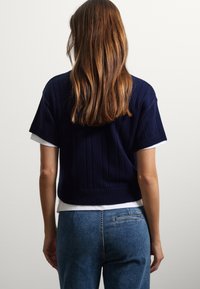 Pull court bleu marine avec texture côtelée et manches courtes, porté sur une chemise blanche, associé à un jean en denim bleu clair.