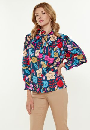 Multifarvet blomsterbluse med bindebånd ved halsen, trekvartærmer og en mørkeblå baggrund med pink, rød og grønne accenter.