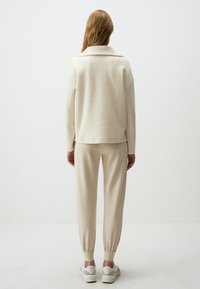 Cremfarbener Strickpullover mit klassischem Kragen, kombiniert mit beigen tapered Jogginghosen und weißen Sneakers. Glatte Textur, lockere Passform.