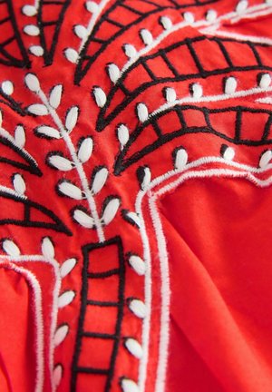 Tissu rouge avec une broderie complexe en noir et blanc représentant des motifs de feuilles et géométriques, avec des points de couture texturés et des couleurs contrastées.