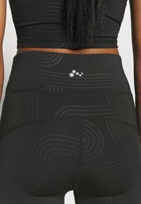 Detailní záběr na osobu v černých legínách s vysokým pasem a černém krátkém topu, ukazující texturované vzory a malé logo na legínách.