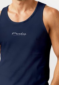 Marinefarbener Tanktop aus glattem Material mit einem weißen Logo-Text "Paradise NEVERLESS" auf der Brust und breiten Schulterträgern.