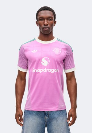 Uomo che indossa una maglia da calcio viola del Manchester United con logo Snapdragon e colletto bianco, abbinata a pantaloncini di jeans blu.