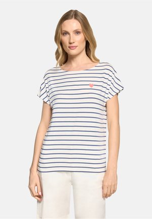 Kobieta ubrana w biało-granatową koszulkę w paski z krótkim rękawem, z małym czerwonym sercem na lewej piersi, w połączeniu z białymi spodniami.