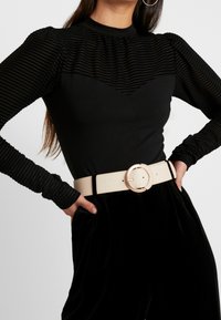 Schwarzes Oberteil mit gerippten Texturen und rundem Ausschnitt, kombiniert mit hoch taillierten schwarzen Samthose und einem breiten beigen Gürtel mit runder Schnalle.