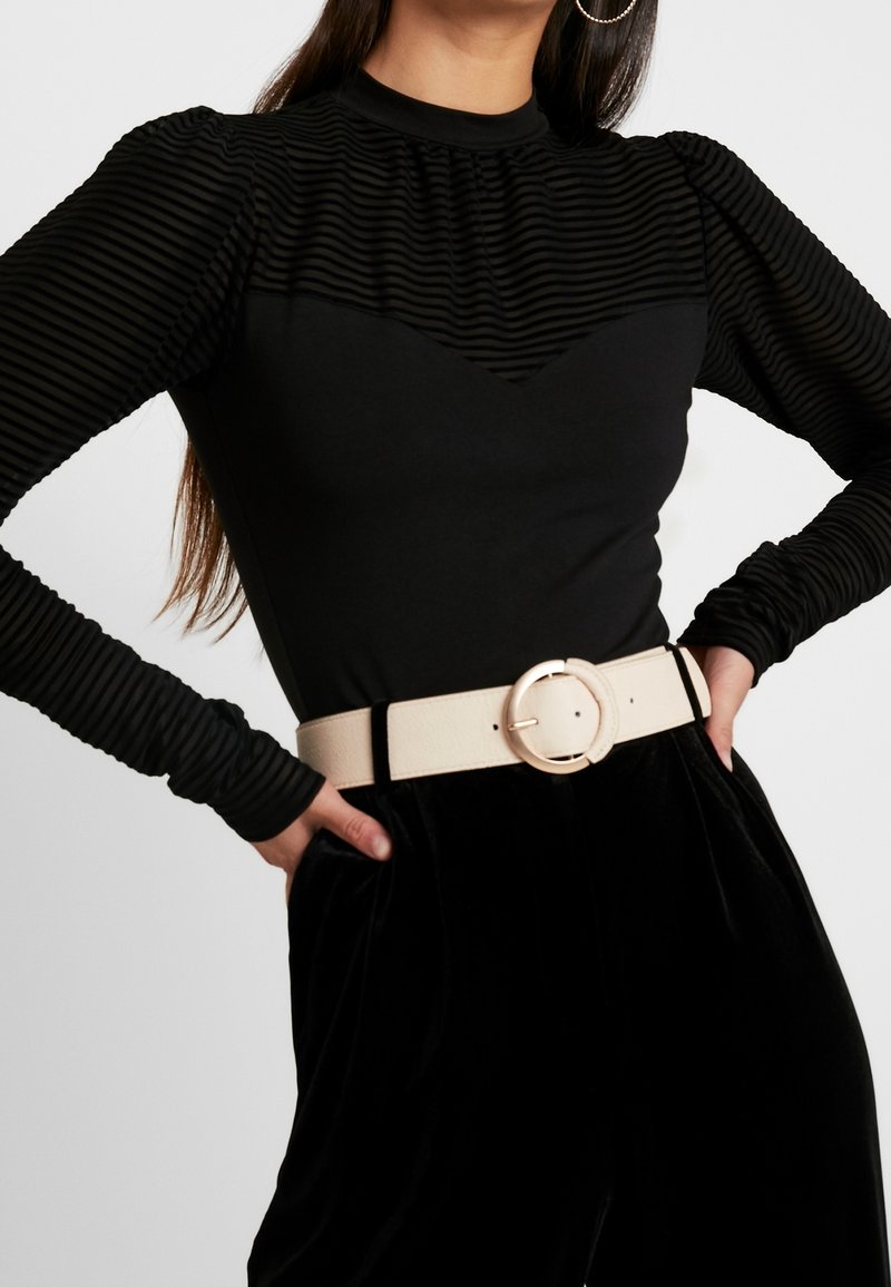 Black top with ribbed textures and a round neckline, paired with high-waisted black velvet pants and a wide beige belt featuring a circular buckle.