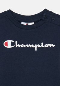 Mornarsko bombažno majico z okroglim izrezom in gumbi na rami. Bele črke "Champion" napisane v teku po sprednjem delu.