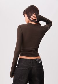 Braunes geripptes Longsleeve-Oberteil mit figurnaher Passform, kombiniert mit schwarzen Jeans, die ein Logo auf dem hinteren Bund und zwei Gesäßtaschen haben.