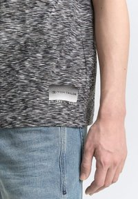 Camiseta gris de manga corta con un patrón texturizado y fluido, y una etiqueta con el logo de Tom Tailor en el dobladillo. Llevada sobre unos jeans de color azul claro.