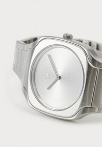 Montre en acier inoxydable argenté avec un cadran carré, finition mates, un cadran minimaliste et un bracelet à maillons, mettant en valeur un simple détail de couronne.