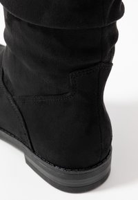 Stivale ankle in suede nero con superficie texturizzata, design curvo e suola piatta. Presenta cuciture sottili lungo le giunture e una base del tacco liscia.