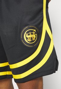 Shorts de sport noirs avec des rayures jaunes et un logo circulaire avec des initiales stylisées. Le tissu est texturé et respirant.