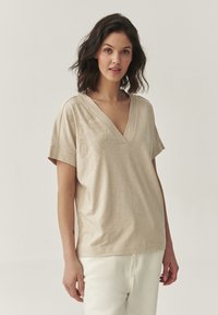 T-shirt v-neck beige à manches courtes, avec une broderie florale sur le côté gauche, fabriqué en tissu léger au toucher lisse.