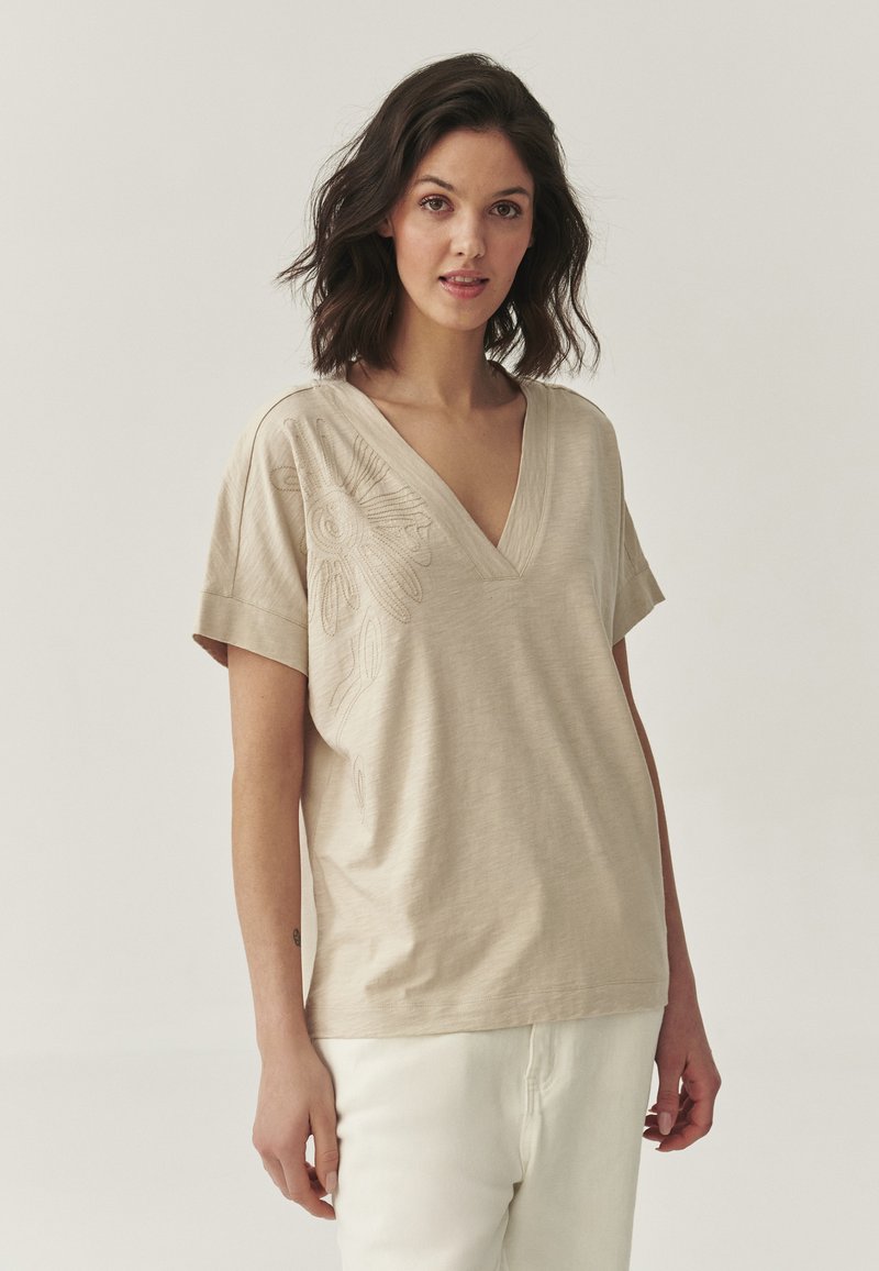 Beige V-Neck-T-Shirt mit kurzen Ärmeln, verziert mit floraler Bestickung auf der linken Seite, gefertigt aus leichtem Stoff mit glatter Textur.