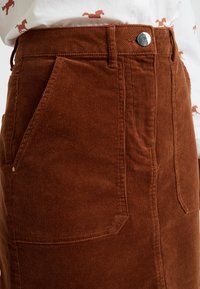 Gonna in velluto a coste marrone con vita aderente, tasche laterali e chiusura con bottone metallico. Il tessuto ha una trama a coste.