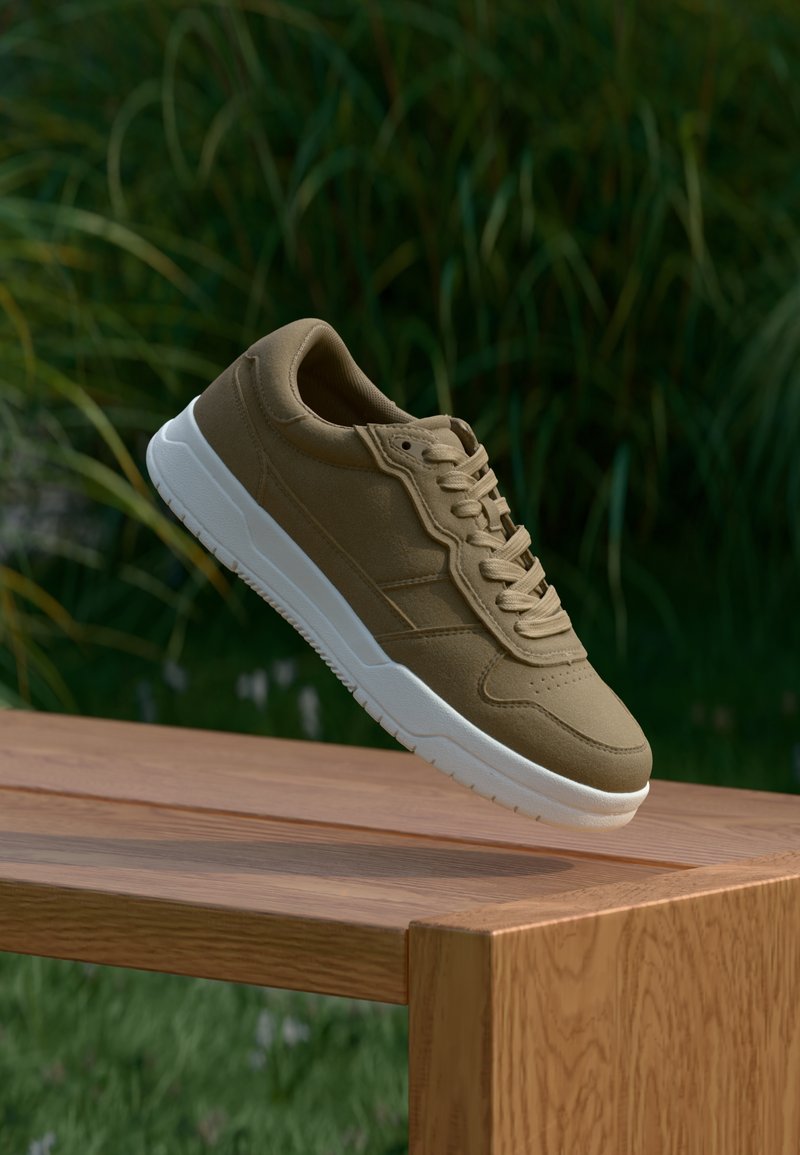 Brune sneaker med hvit såle som svever over en trebenk, gress og høye planter i den uskarpe bakgrunnen.