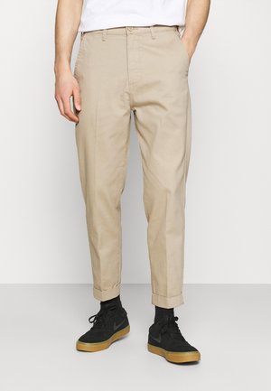 Mężczyzna ubrany w beżowe spodnie z mankietami, czarne skarpetki i czarne trampki z brązowymi podeszwami, stojący na jednolitym jasnym tle.