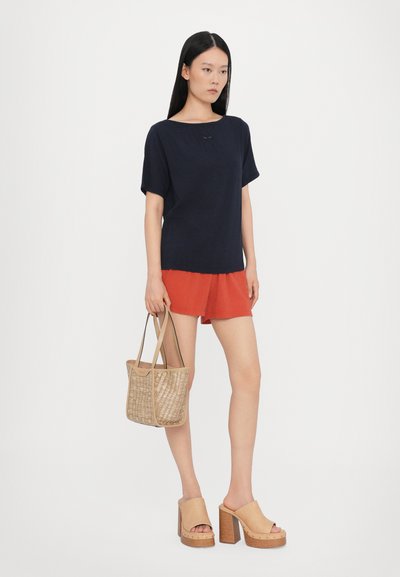 Top in cotone blu navy a maniche corte, pantaloni corti rossi, sandali beige con tacco spesso e borsa a tracolla intrecciata. Design semplice con vestibilità rilassata e texture liscia.