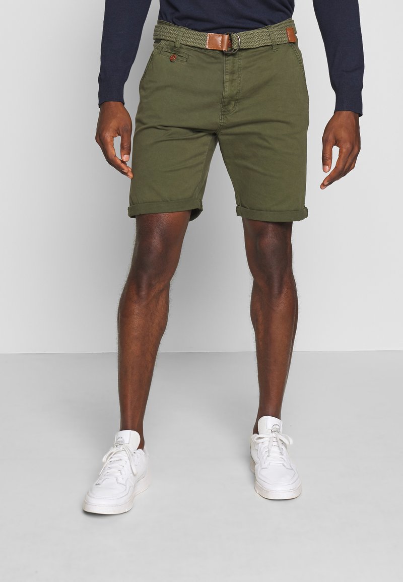 Shorts en coton vert olive avec revers, présentant une ceinture tressée avec un accent en cuir marron et des poches avant. Des baskets blanches complètent le look.