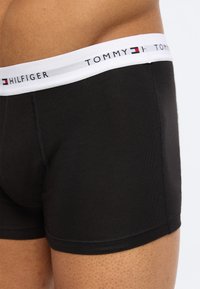 Κοντινό πλάνο ενός ατόμου που φοράει μαύρα ανδρικά εσώρουχα Tommy Hilfiger με άσπρη ζώνη μέσης που φέρει το λογότυπο και το έμβλημα της σημαίας της μάρκας.