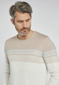 Lekki sweter z dzianiny w kolorze beżowym i białym, z niebieskimi i jasnobrązowymi paskami na wysokości klatki piersiowej. Posiada klasyczny okrągły dekolt i prążkowane mankiety.