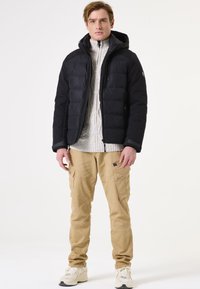 Schwarze Daunenjacke mit Kapuze, mit Reißverschlusstaschen vorne und einem strukturierten grauen Strickpullover darunter. Sandfarbene Cargo-Hosen, weiße Sneaker.