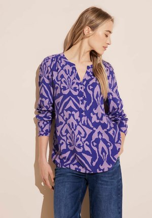 Paarse blouse met lange mouwen en een ingewikkeld lavendelpuntpatroon, V-hals en knoopdetails. Gecombineerd met een blauwe spijkerbroek. Zachte, lichte stof.