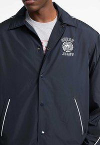 Marineblaue, leichte Jacke mit klassischem Kragen, Druckknöpfen, gesticktem Logo und weißen Akzenten an den Taschen und Ärmeln.