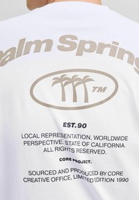 Hvid bomulds t-shirt med "Palm Springs" i beige, med palme grafisk design, sort tekst og forskellige mærker på bagsiden.