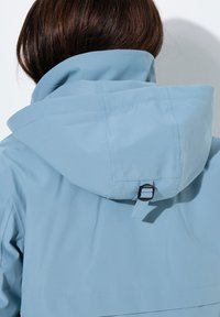Chaqueta impermeable azul claro con capucha acolchada y correa trasera ajustable. Textura suave y diseño elegante con costuras mínimas.