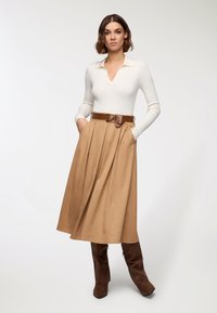 Maglietta bianca a costine a maniche lunghe con scollo a V, abbinata a una gonna plissettata beige e una cintura in pelle marrone. La modella indossa stivali marroni sopra il ginocchio.