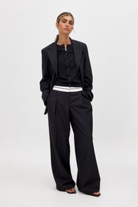 Schwarze Oversized-Blazer mit strukturiertem Design, kombiniert mit einer taillierten schwarzen Hemdbluse und weiten Hosen, die einen weißen Taillengürtel haben.