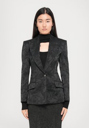 Woman with long black hair wearing a patterned black blazer over a black knit top and sparkly dark skirt, standing against a plain background.