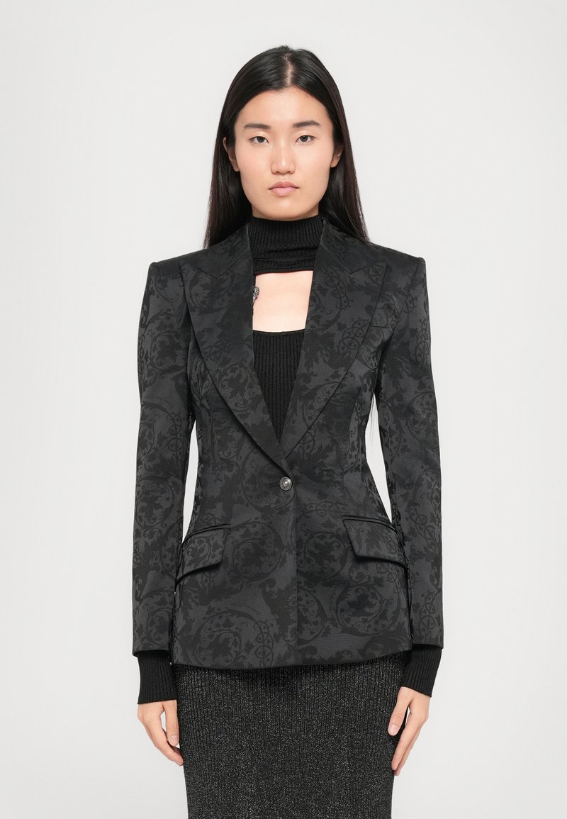 Woman with long black hair wearing a patterned black blazer over a black knit top and sparkly dark skirt, standing against a plain background.