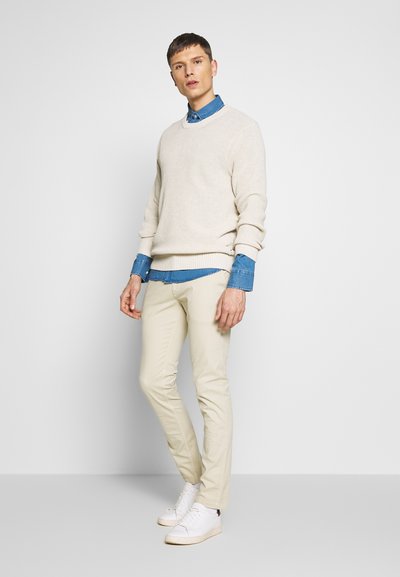Beige gestrickter Pullover über einem blauen Jeanshemd, kombiniert mit hellbeigen Hosen und weißen Sneakers. Lässiger Lagenlook mit strukturiertem Finish.