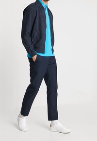 Marynarka bombowa w kolorze granatowym, jasnoniebieska koszulka polo, dopasowane granatowe spodnie i białe sneakersy. Gładki materiał, sportowy krój i wyraźne linie w całym kroju.