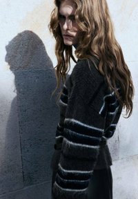 Sivi prugasti pulover z mehkim materialom, s belimi in mornarsko modrimi detajli, nosi oseba proti ozadju iz kamna.