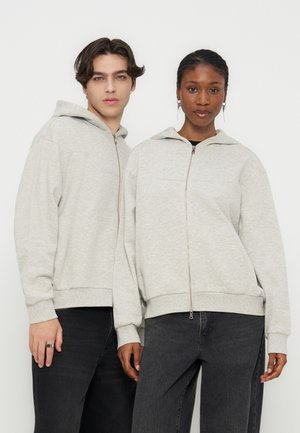 Deux jeunes adultes portant des sweats à capuche gris clair zippés et des pantalons foncés, debout près l’un de l’autre sur un fond blanc uni.