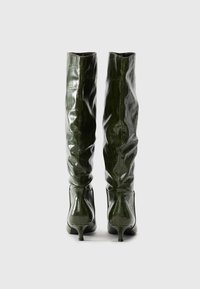 Un paio di stivali alti lucidi di un verde scuro con una texture a coccodrillo, visti da dietro su uno sfondo chiaro.