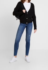 Person wearing black buttoned cardigan, white shirt, blue skinny jeans, white sneakers, holding black shoulder bag on a white background.