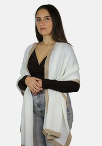 Biały szal ze złotym metalicznym obszyciem, narzucony na brązową bluzkę. Tkanina wydaje się miękka o gładkiej fakturze, doskonale uzupełniająca casualowy strój.