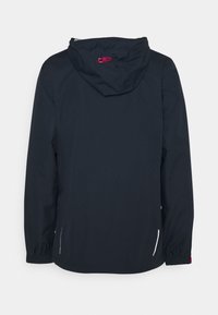Veste imperméable bleu marine avec capuche. Dispose de poignets élastiques, de poches latérales avec fermetures éclair blanches et d'un logo CMP rouge au niveau du col.