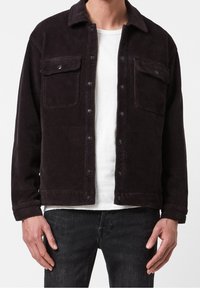 Man i en svart manchesterjacka med tryckknappar över en vit t-shirt och mörk tvättade jeans, står med avslappnade händer.