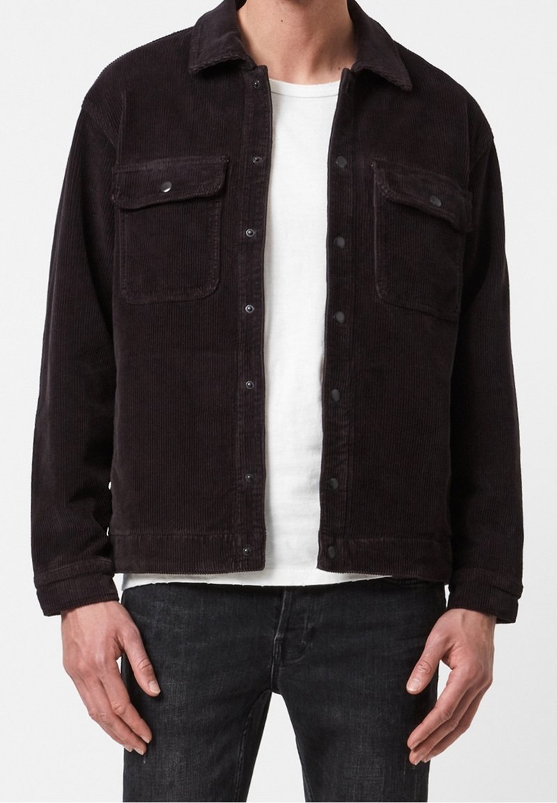 Man i en svart manchesterjacka med tryckknappar över en vit t-shirt och mörk tvättade jeans, står med avslappnade händer.