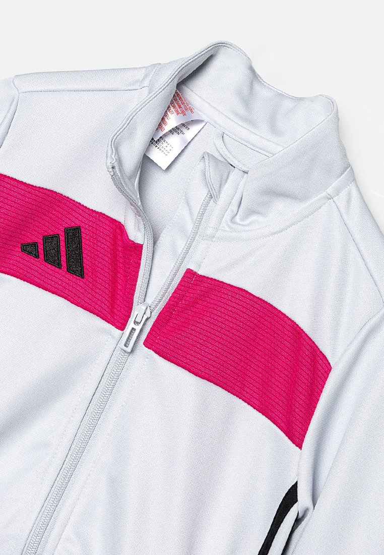 Giacca sportiva grigia chiara con zip frontale, caratterizzata da ampie strisce rosa e nere e un logo nero sul lato sinistro. Texture liscia.