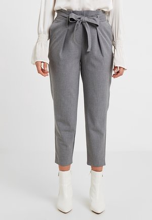 Vrouw die grijze broek met hoge taille en taps toelopende pijpen draagt, met een stoffen riem geknoopt in een strik, gecombineerd met witte enkellaarsjes en een witte blouse met gerimpelde manchetten.