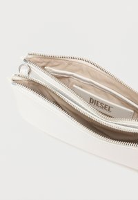 Λευκό δερμάτινο πορτοφόλι με πολλαπλές θήκες με φερμουάρ, ασημένια εξαρτήματα, λείο εξωτερικό και εσωτερική επένδυση από μπεζ ύφασμα με ετικέτα λογότυπου.