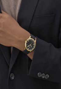 Black wristwatch with gold-tone case and Roman numeral dial, featuring a silicone strap. Worn on a wrist, complemented by a dark blazer.