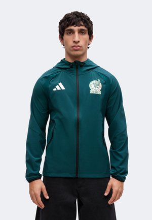 Homme portant une veste vert foncé zippée avec le logo de l’équipe de football du Mexique et des bandes Adidas, debout sur un fond clair.